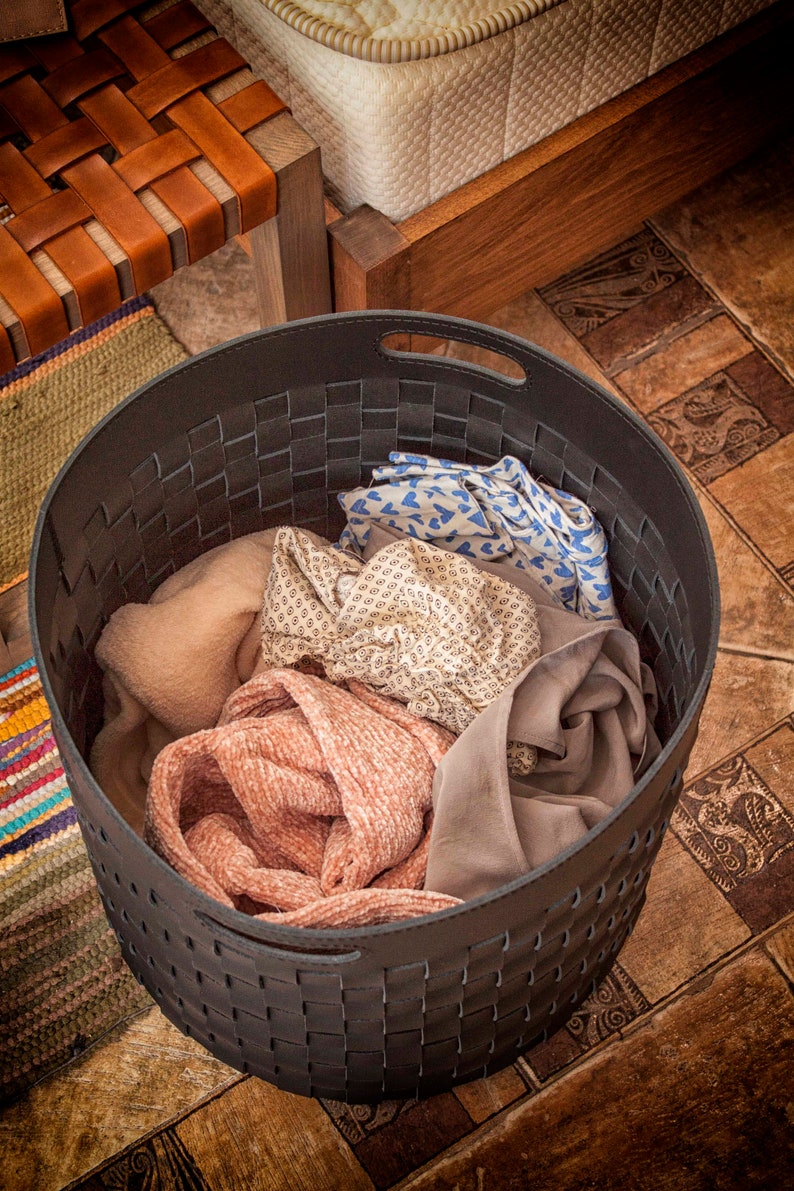 Puede incluir: Una cesta de almacenamiento redonda de tejido gris oscuro con asas integradas, llena de una variedad de textiles plegados que incluyen una tela rosa texturizada, una tela beige estampada, una tela azul con corazones y una pieza de tela gris lisa.