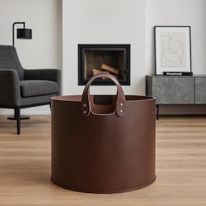 May include: A brown leather log carrier with a rounded base and a handle. The cylindrical carrier has a handle attached with rivets. The interior is a lighter shade of brown. The carrier is designed for indoor use.