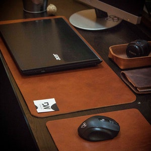 May include: A desk setup with a black laptop on a brown leather desk mat. A black mouse sits on a smaller matching mat. A business card and a camera lens are also present. The desk is made of dark wood.