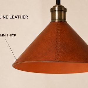 May include: A conical pendant lamp shade crafted from brown genuine leather, 3-3.5 mm thick, featuring visible cross-stitching along the side and a metallic brass-toned fixture at the top. The interior of the shade is a smooth, lighter orange tone.