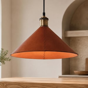 May include: A conical pendant light fixture featuring a smooth, rust-colored leather shade with visible zigzag stitching along the side. The lamp is suspended by a black cord and includes a metallic brass-toned hardware fitting at the top.