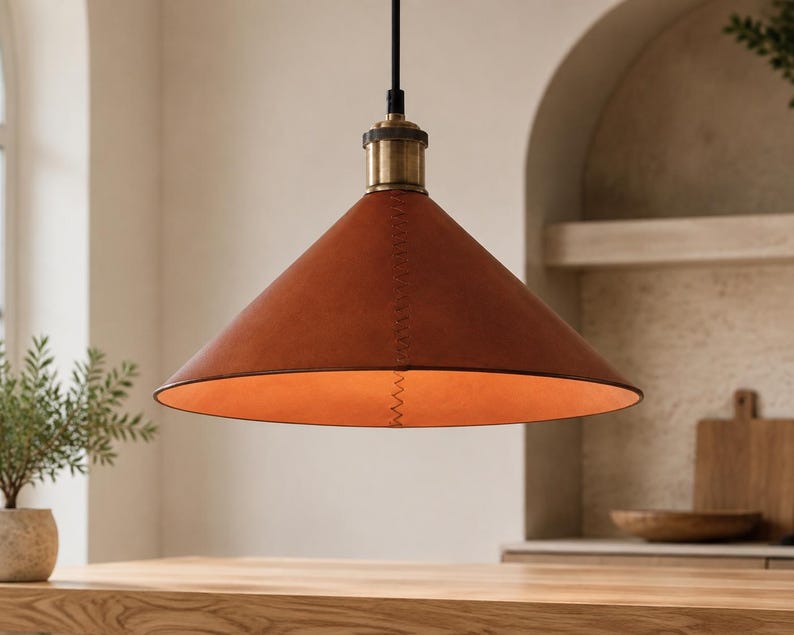May include: A conical pendant light fixture featuring a smooth, rust-colored leather shade with visible zigzag stitching along the side. The lamp is suspended by a black cord and includes a metallic brass-toned hardware fitting at the top.