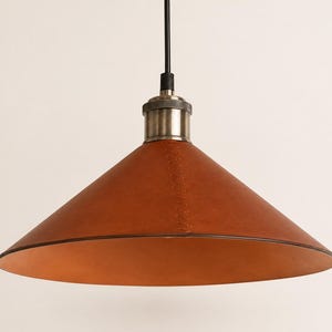 May include: A conical pendant light fixture featuring a smooth, tan leather shade with visible vertical stitching. The shade is attached to a metallic, brushed-nickel finish socket and a black cord, creating a clean, functional lighting design.