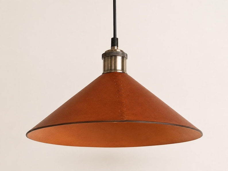 May include: A conical pendant light fixture featuring a smooth, tan leather shade with visible vertical stitching. The shade is attached to a metallic, brushed-nickel finish socket and a black cord, creating a clean, functional lighting design.