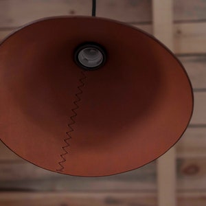 May include: A conical pendant lamp shade in a matte terracotta brown finish. The shade features a prominent vertical seam joined by dark brown zigzag stitching that runs from the central light bulb socket down to the wide circular bottom edge.