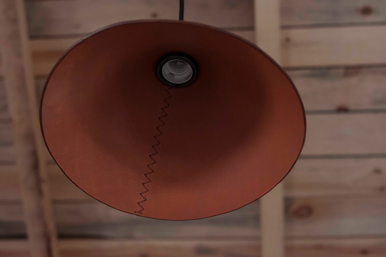 May include: A conical pendant lamp shade in a matte terracotta brown finish. The shade features a prominent vertical seam joined by dark brown zigzag stitching that runs from the central light bulb socket down to the wide circular bottom edge.