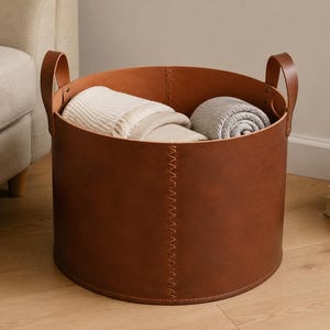 Puede incluir: Una cesta de almacenamiento redonda de cuero marrón con dos asas resistentes y una costura vertical visible. El interior de la cesta contiene dos mantas dobladas, una de color crema y la otra en un tono gris claro.