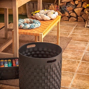 Puede incluir: Una cesta de almacenamiento cil&iacute;ndrica de color gris oscuro con un patr&oacute;n exterior trenzado hecho de tiras anchas y planas. La cesta tiene un borde s&oacute;lido con dos asas recortadas integradas en los lados opuestos, con un dise&ntilde;o abierto.