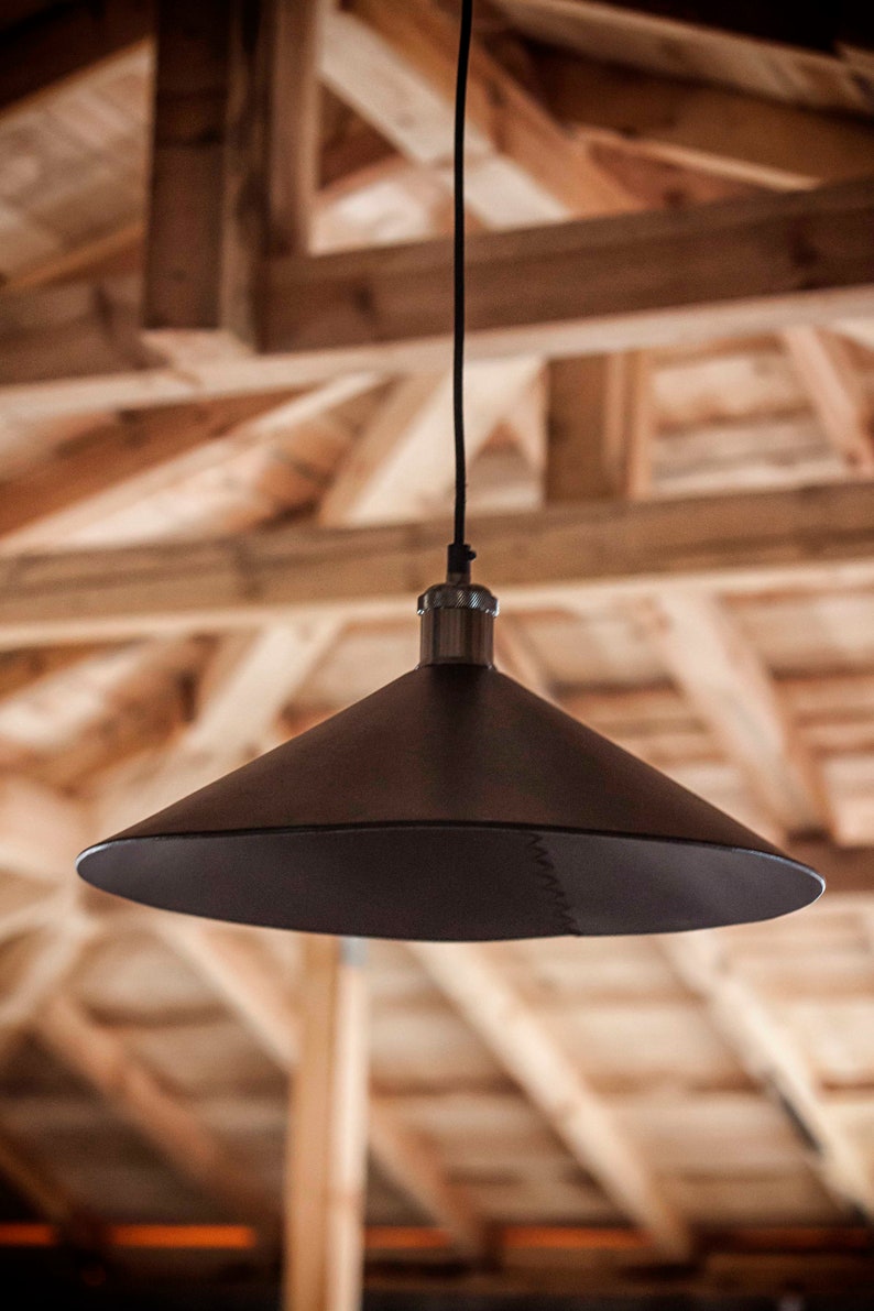 May include: A matte black conical pendant light fixture suspended by a thin black cord. The shade features a visible stitched seam running vertically along one side, connecting to a metallic silver-toned hardware fitting at the top center.