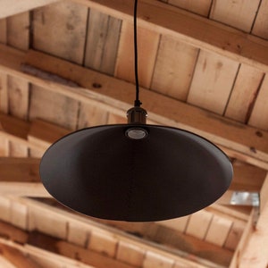 May include: A matte black pendant light fixture featuring a wide, conical shade suspended by a thin black cord. The lamp includes a metallic socket housing at the top center, with a visible seam running vertically down the surface of the shade.