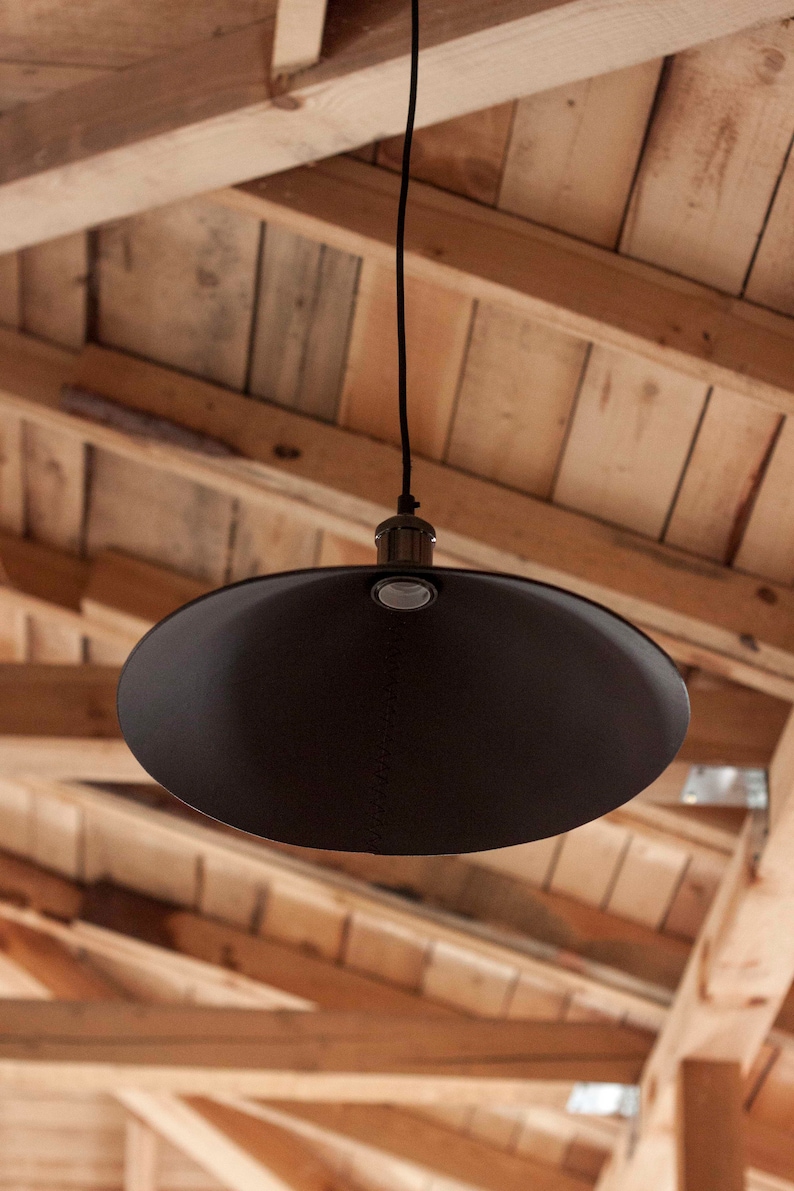 May include: A matte black pendant light fixture featuring a wide, conical shade suspended by a thin black cord. The lamp includes a metallic socket housing at the top center, with a visible seam running vertically down the surface of the shade.