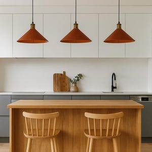 May include: Three conical terracotta-colored pendant lights with black cords hang above a light oak kitchen island. Two light oak wooden spindle-back bar stools are positioned in front of the island, which features a smooth, flat countertop surface.