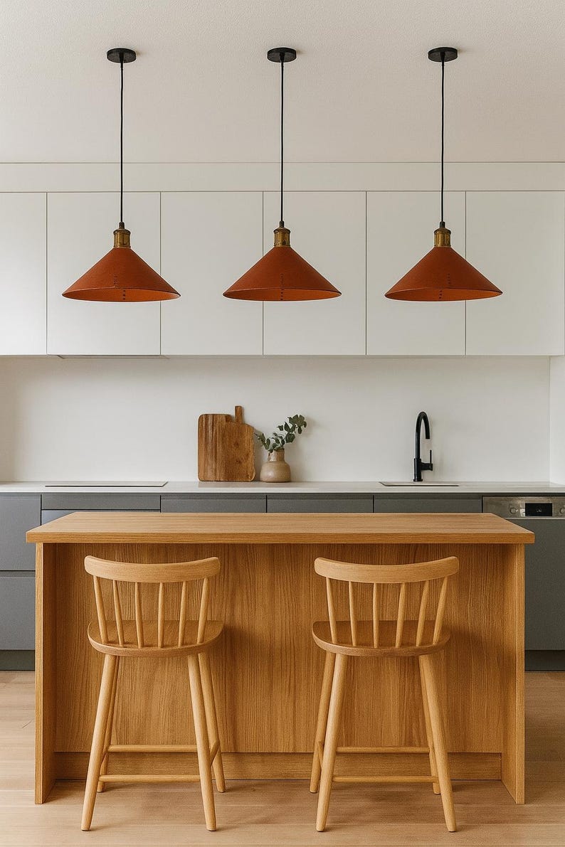 May include: Three conical terracotta-colored pendant lights with black cords hang above a light oak kitchen island. Two light oak wooden spindle-back bar stools are positioned in front of the island, which features a smooth, flat countertop surface.