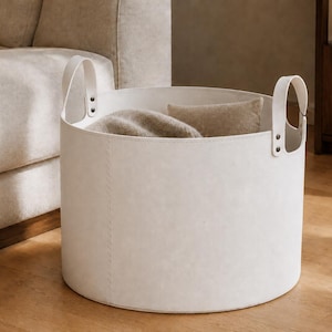 May include: A round, off-white storage basket with a smooth, matte finish and two vertical handles attached by metal rivets. The basket has visible stitched seams along the side and contains a soft, textured beige fabric item inside.