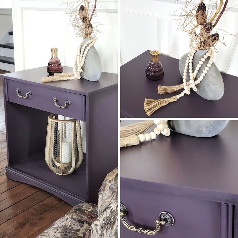 May include: A purple painted side table with a shelf, two drawers, and decorative accents. The table is decorated with a white beaded garland, a candle in a wire cage, and a glass bottle with a stopper.