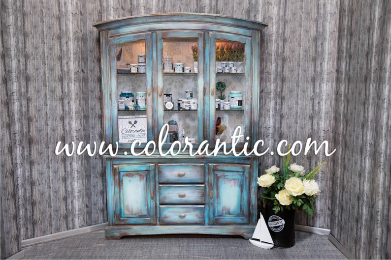 May include: A distressed turquoise blue hutch with glass doors and drawers. The hutch is filled with various jars and bottles of paint. The website www.colorantic.com is displayed on the front of the hutch.