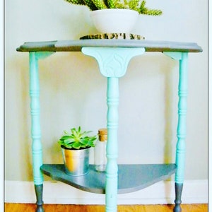 May include: A small, gray and teal corner table with a shelf. The table has three legs and a decorative top. A small succulent plant is on the shelf.