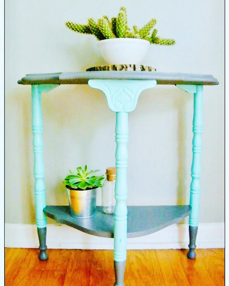 May include: A small, gray and teal corner table with a shelf. The table has three legs and a decorative top. A small succulent plant is on the shelf.