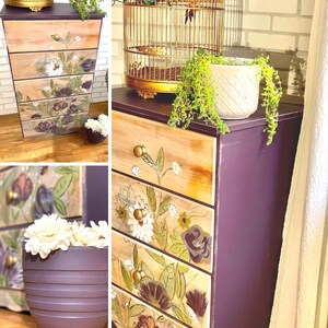 May include: A five-drawer chest of drawers with a purple painted finish and floral designs on the drawers. The chest has gold knobs and is sitting on a wooden floor.