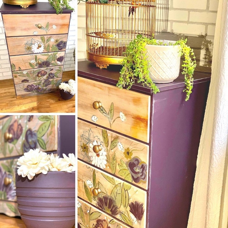 May include: A five-drawer chest of drawers with a purple painted finish and floral designs on the drawers. The chest has gold knobs and is sitting on a wooden floor.