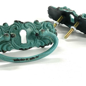 May include: Two turquoise blue metal drawer pulls with ornate designs and keyhole cutouts. The pulls have a distressed finish and are attached to screws.