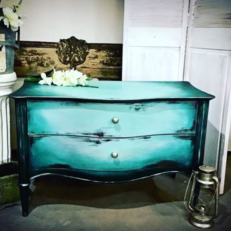 Op de afbeelding: Een dressoir met twee laden, een gebogen voorzijde en verweerde teal verf. Het dressoir heeft zwarte accenten en zilveren ladegrepen.