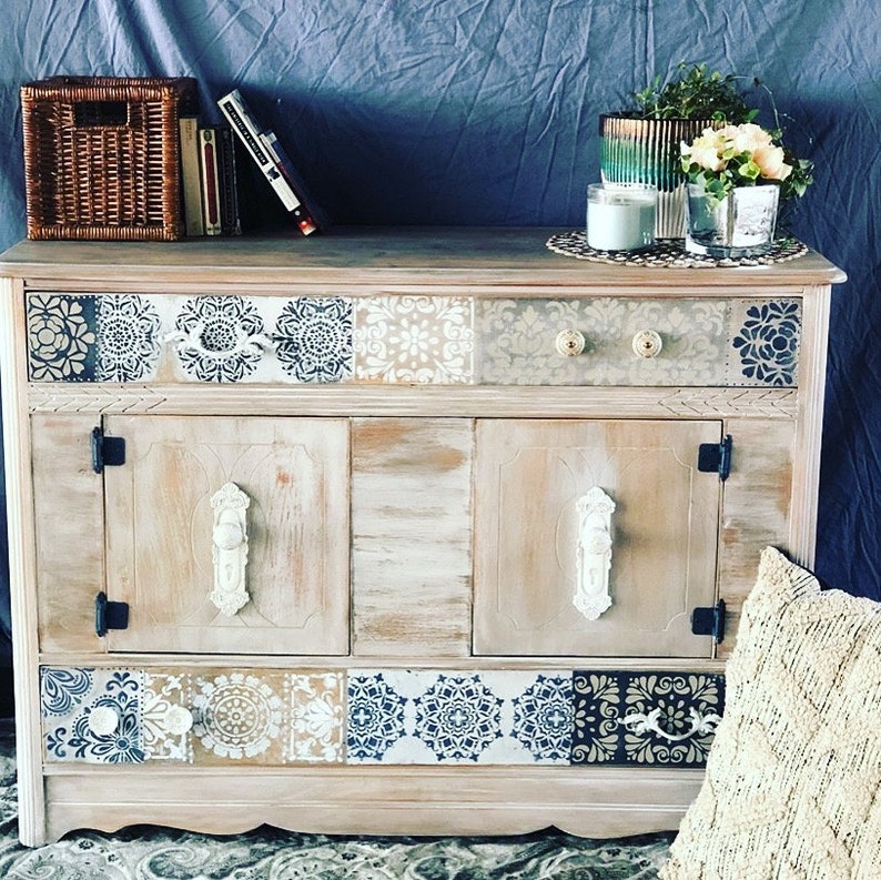 May include: A distressed white wooden cabinet with two doors and a drawer. The cabinet has a decorative tile pattern on the top and bottom. The cabinet is decorated with black metal hardware and white ceramic knobs.