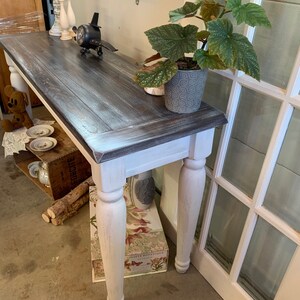 Op de afbeelding: Een wit geverfde consoletafel met een donkerhouten blad. De tafel heeft gedraaide poten en is versierd met een klein vliegtuig, kaarsen en een plant in een pot.