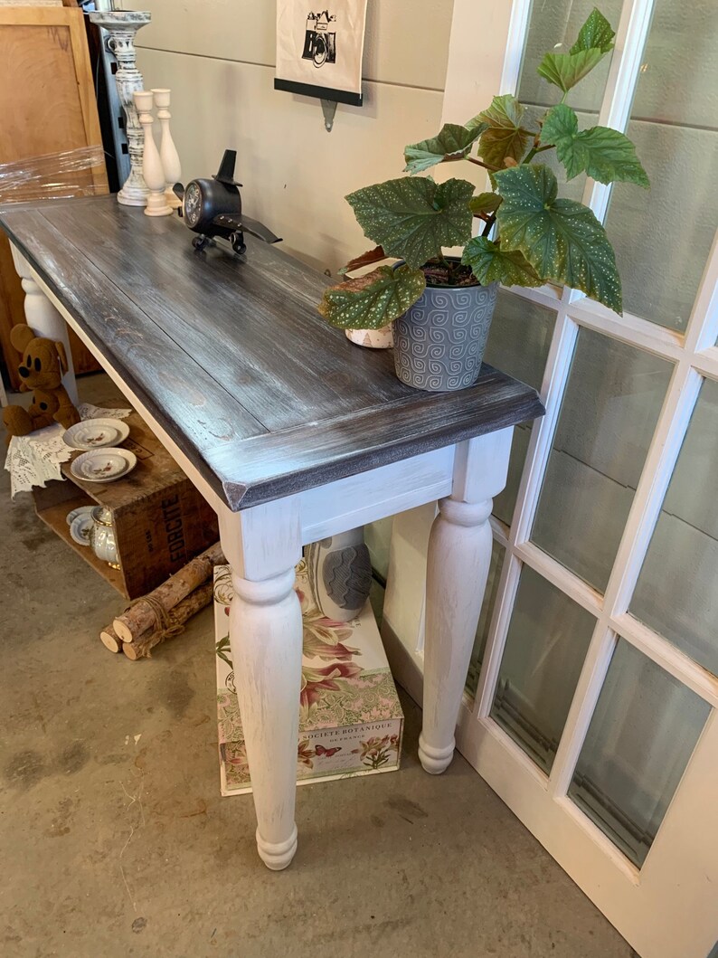 Op de afbeelding: Een wit geverfde consoletafel met een donkerhouten blad. De tafel heeft gedraaide poten en is versierd met een klein vliegtuig, kaarsen en een plant in een pot.