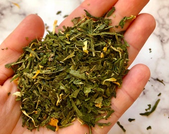 May include: A close-up of a hand holding a loose leaf green tea blend with yellow flower petals.