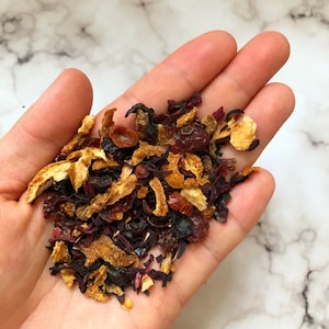 May include: A close-up of a hand holding a mixture of dried fruit, orange peel, and herbs. The mixture is a blend of red, orange, and brown colors.