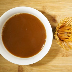 May include: A white ceramic bowl filled with brown liquid, likely coffee or tea, and a bamboo whisk for whisking matcha tea.