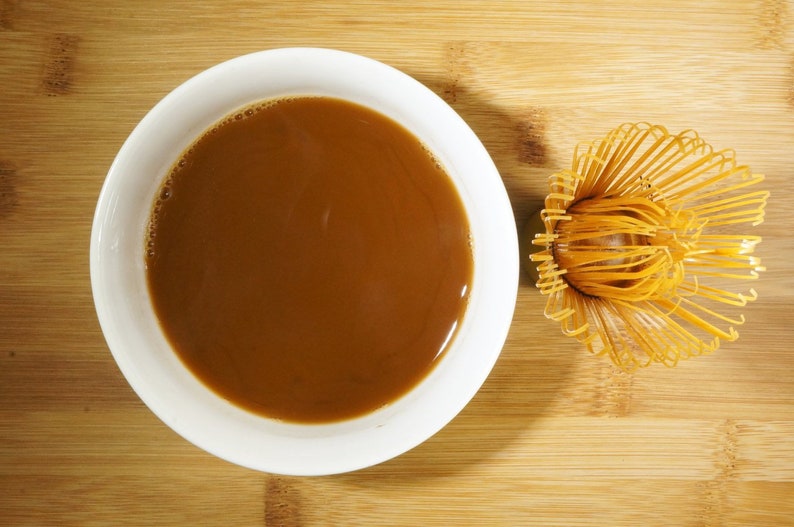 May include: A white ceramic bowl filled with brown liquid, likely coffee or tea, and a bamboo whisk for whisking matcha tea.