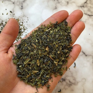 May include: A close-up of a hand holding a pile of loose leaf green tea. The tea is a blend of green and black tea leaves.