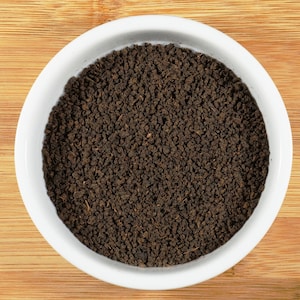 May include: A white bowl filled with loose leaf black tea. The tea is a dark brown color and has a rough texture.