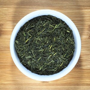 May include: A white bowl filled with loose green tea leaves. The bowl is sitting on a wooden surface.