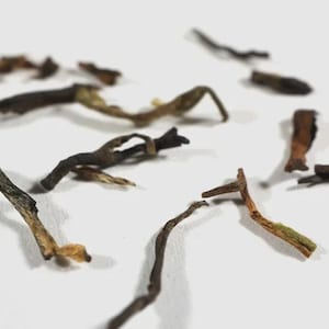 May include: A collection of loose-leaf tea scattered across a surface, featuring dried, twisted tea leaves in shades of dark brown, black, and light tan, with some pieces showing thin, elongated stems and curled, textured foliage.