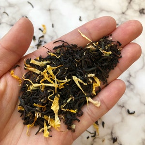May include: A close-up of a hand holding a blend of black tea leaves and dried mango pieces.