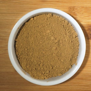 May include: A white ceramic bowl filled with a light brown powder. The powder is evenly distributed in the bowl and appears to be a fine texture.
