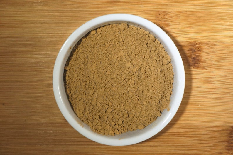 May include: A white ceramic bowl filled with a light brown powder. The powder is evenly distributed in the bowl and appears to be a fine texture.