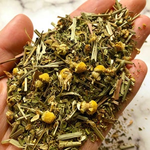 May include: A handful of loose herbal tea blend, featuring dried chamomile flowers and green leaves. The tea blend includes yellow chamomile blossoms and long green grass-like herbs. The tea is a mix of green and yellow hues.