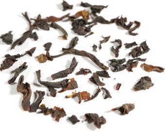 May include: Loose black tea leaves scattered on a white surface. The leaves are dry and have a dark brown color.