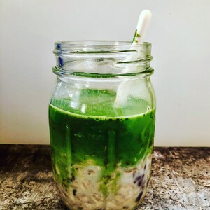 Puede incluir: Un tarro de vidrio lleno de un batido de matcha verde en capas sobre una base cremosa blanca con semillas negras y blancas. Una cuchara de pl&aacute;stico blanca sobresale de la parte superior del tarro.