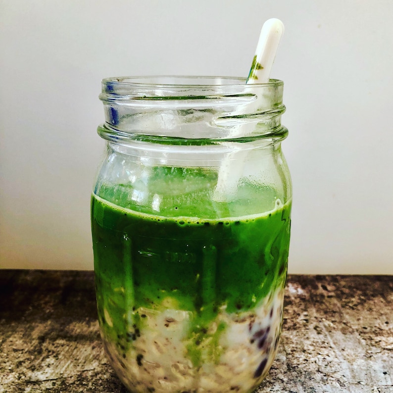 Puede incluir: Un tarro de vidrio lleno de un batido de matcha verde en capas sobre una base cremosa blanca con semillas negras y blancas. Una cuchara de pl&aacute;stico blanca sobresale de la parte superior del tarro.