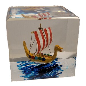 May include: A miniature Viking ship with a red and white striped sail and a yellow hull is encased in a clear resin cube. The base of the cube features blue wave-like patterns, creating a nautical theme. The ship is a decorative item.