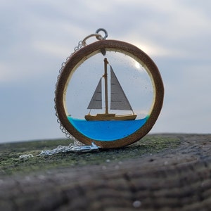 May include: A round pendant necklace featuring a miniature sailboat encased in clear resin. The sailboat has a beige hull and white sails, set against a blue water background. The pendant is framed by a wooden ring and hangs on a silver chain.