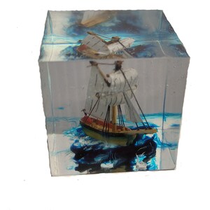 May include: A small wooden sailboat model encased in clear resin with blue and white swirling accents. The sailboat has white sails and a red hull.