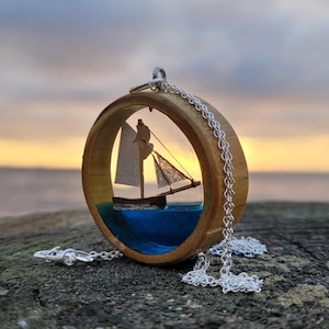 May include: A wooden pendant necklace with a clear resin circle containing a miniature sailboat. The sailboat is white with a brown hull and is set against a blue resin background. The pendant is attached to a silver chain.