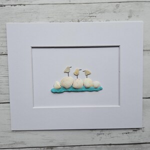 May include: A framed artwork depicting three small bird figures made from seashells. The birds are positioned on a cluster of white seashells and a strip of turquoise material, set against a white background. The artwork is in a white mount and frame.