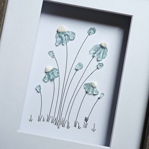 May include: Framed artwork with a floral design. Light blue glass pieces form flowers with white centres on a white background. The artwork is set within a gray wooden frame, creating a delicate and artistic piece.